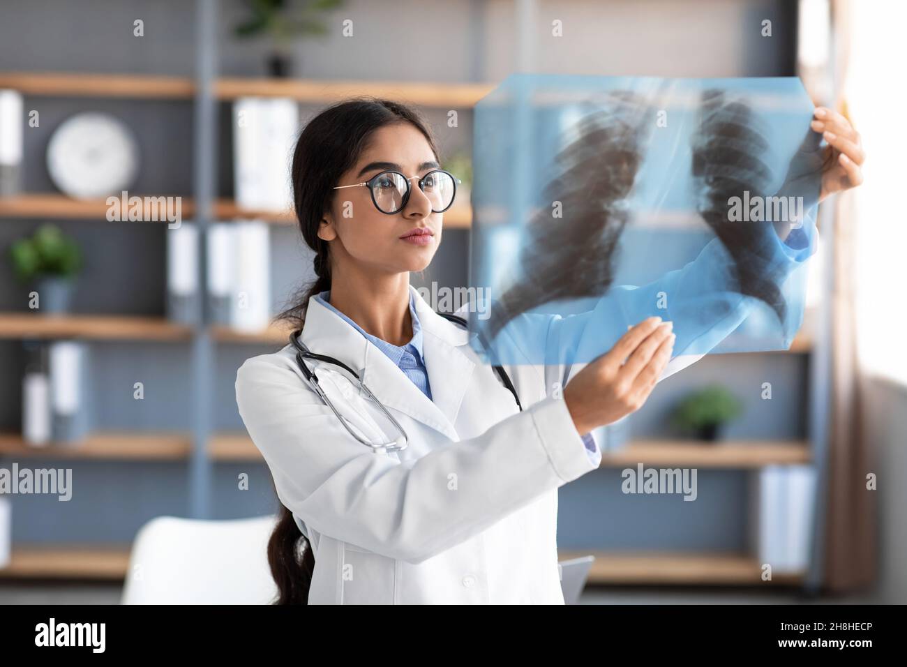 Occupé sérieux millénaires jolie femme indienne médecin en manteau blanc et des lunettes regardant les poumons x-ray dans la clinique Banque D'Images