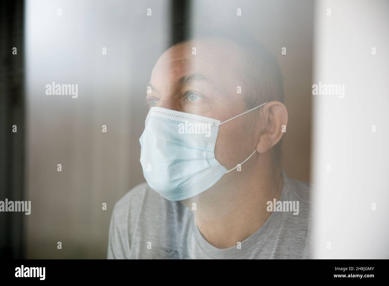 Homme avec un masque de visage assis dans une quarantaine à la maison et regardant par la fenêtre. Banque D'Images