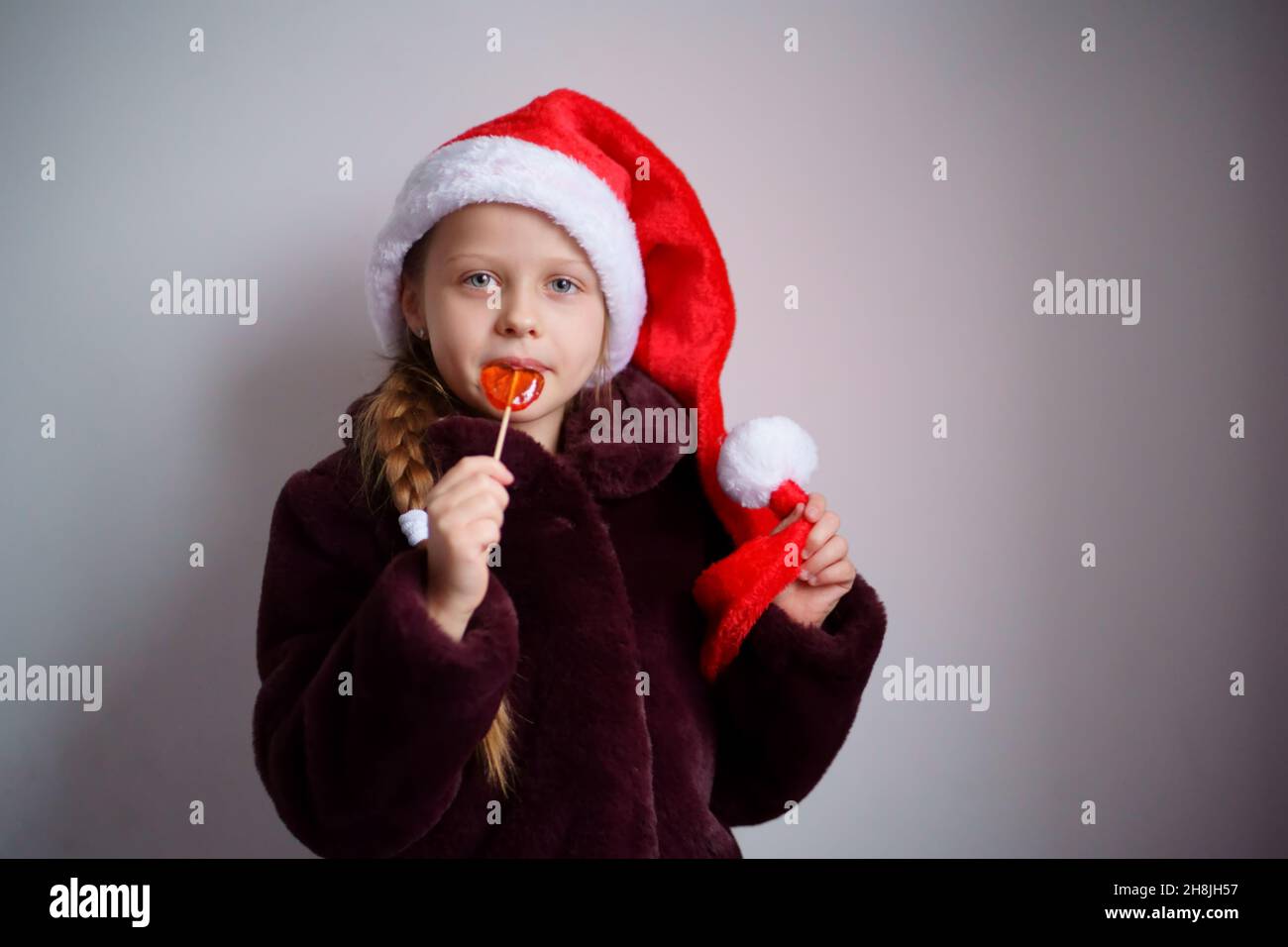 Une fille de Noël blanche en casquette rouge et fourrure du Père Noël liche un lollipop contre un fond clair. Banque D'Images