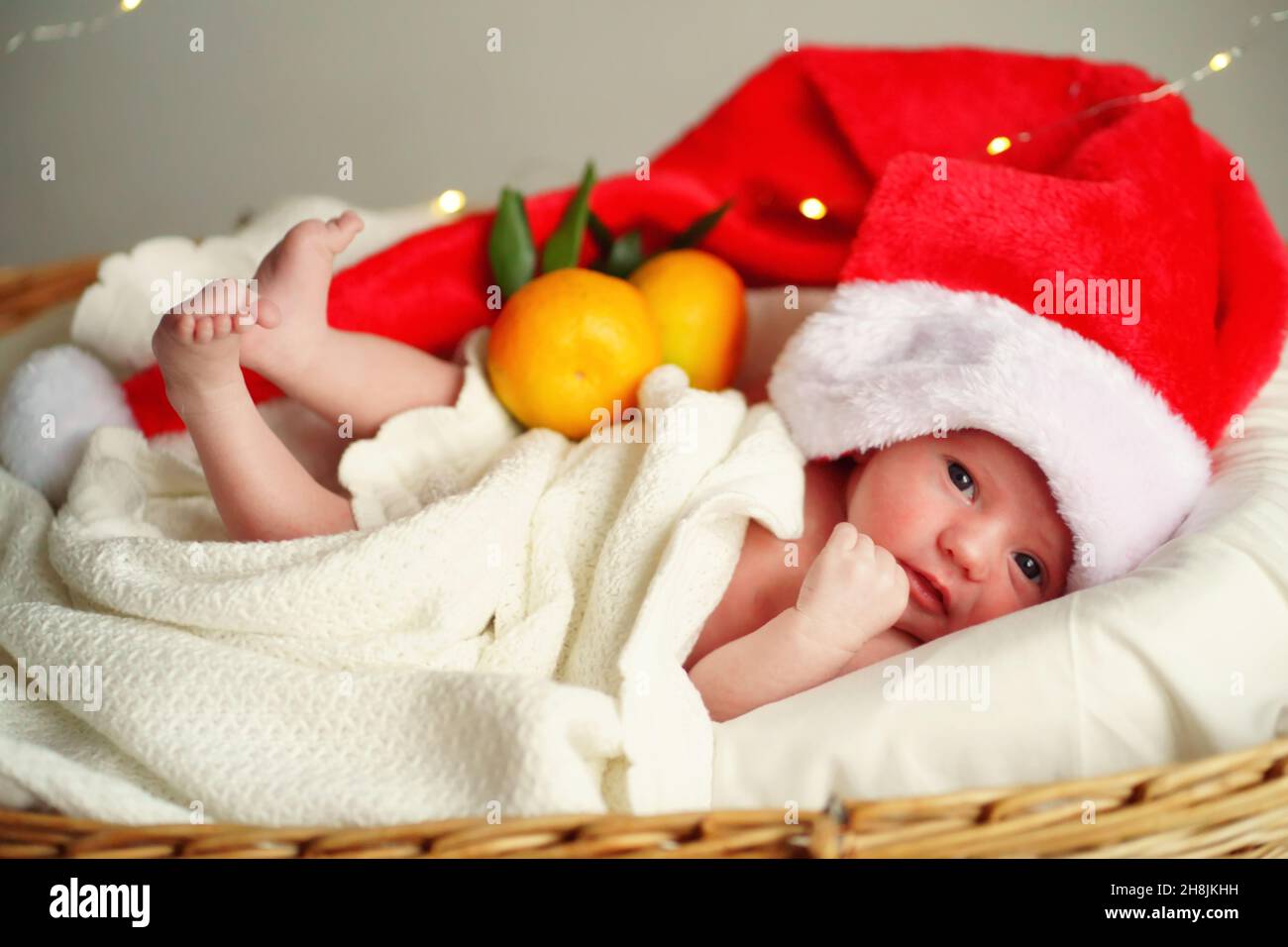 Le nouveau-né caucasien dans la casquette de Noël du Père Noël se trouve dans un panier avec des mandarines sous une couverture blanche, des lumières brûlantes contre l'arrière-plan. Banque D'Images