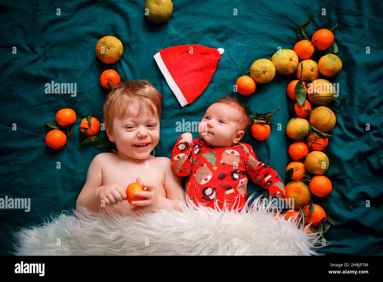 Le frère aîné est avec un nouveau-né de Noël sur un lit vert confortable, à côté du chapeau du Père Noël et le numéro un des mandarines.Les bébés le sont Banque D'Images