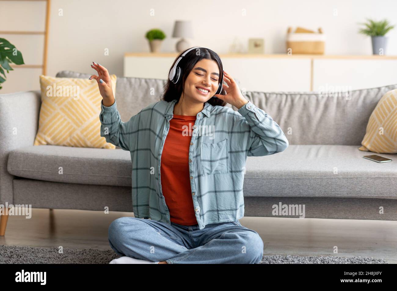 Jeune femme arabe positive assise sur le sol avec un casque, écoutant de la musique ou un livre audio à la maison Banque D'Images