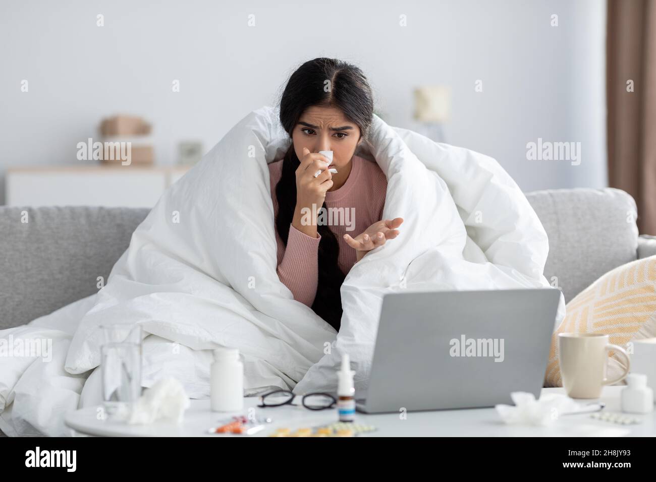 Jeune femme hindoue malheureuse enveloppée dans une couverture, souffre de la grippe, sur le canapé, le nez de souffle regarde l'ordinateur Banque D'Images