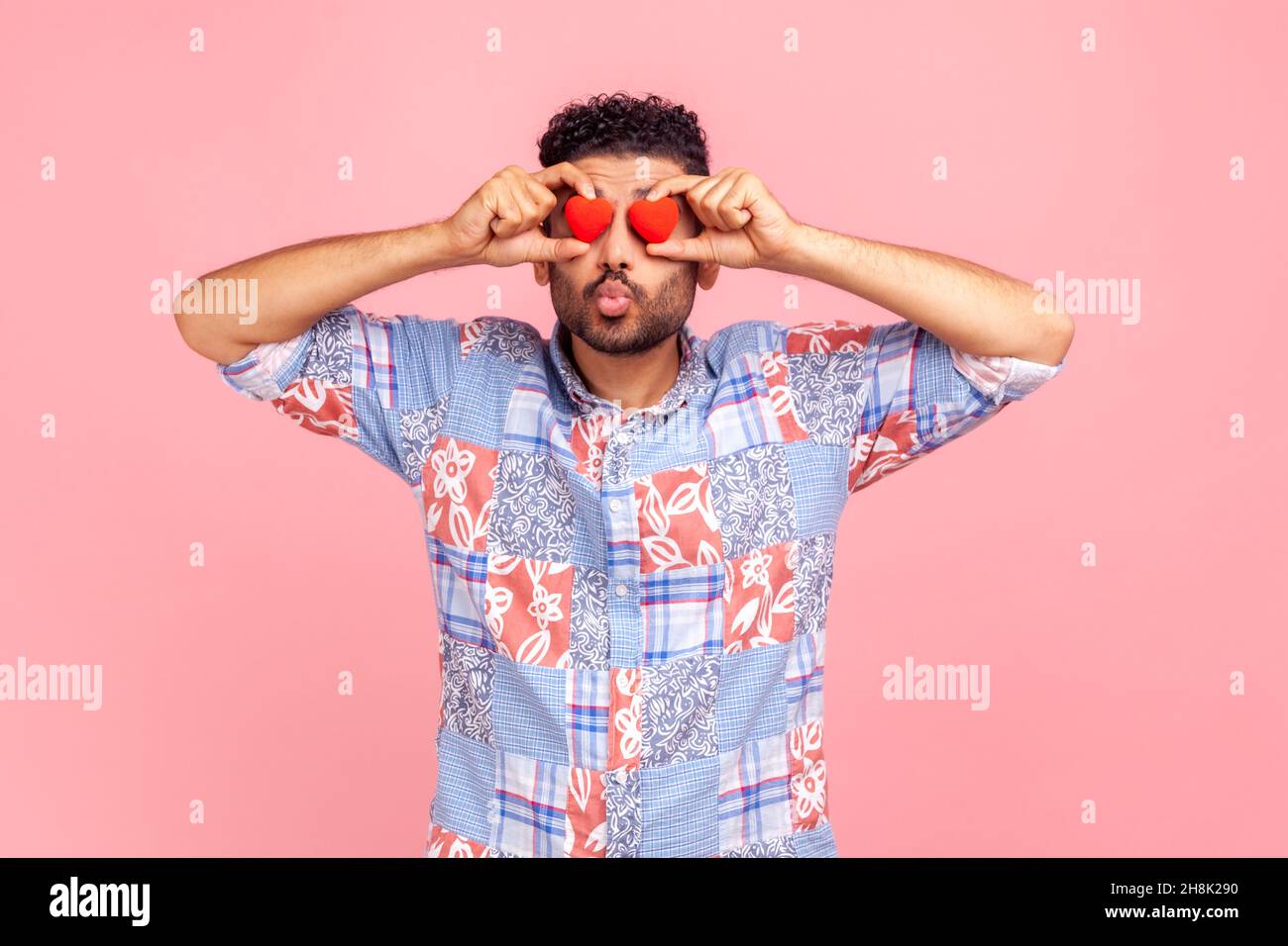 Jeune adulte barbu beau homme avec des lèvres pout, faisant un geste de baiser, couvrant les yeux avec de petits coeurs rouges, portant bleu décontracté style chemise.Studio d'intérieur isolé sur fond rose. Banque D'Images