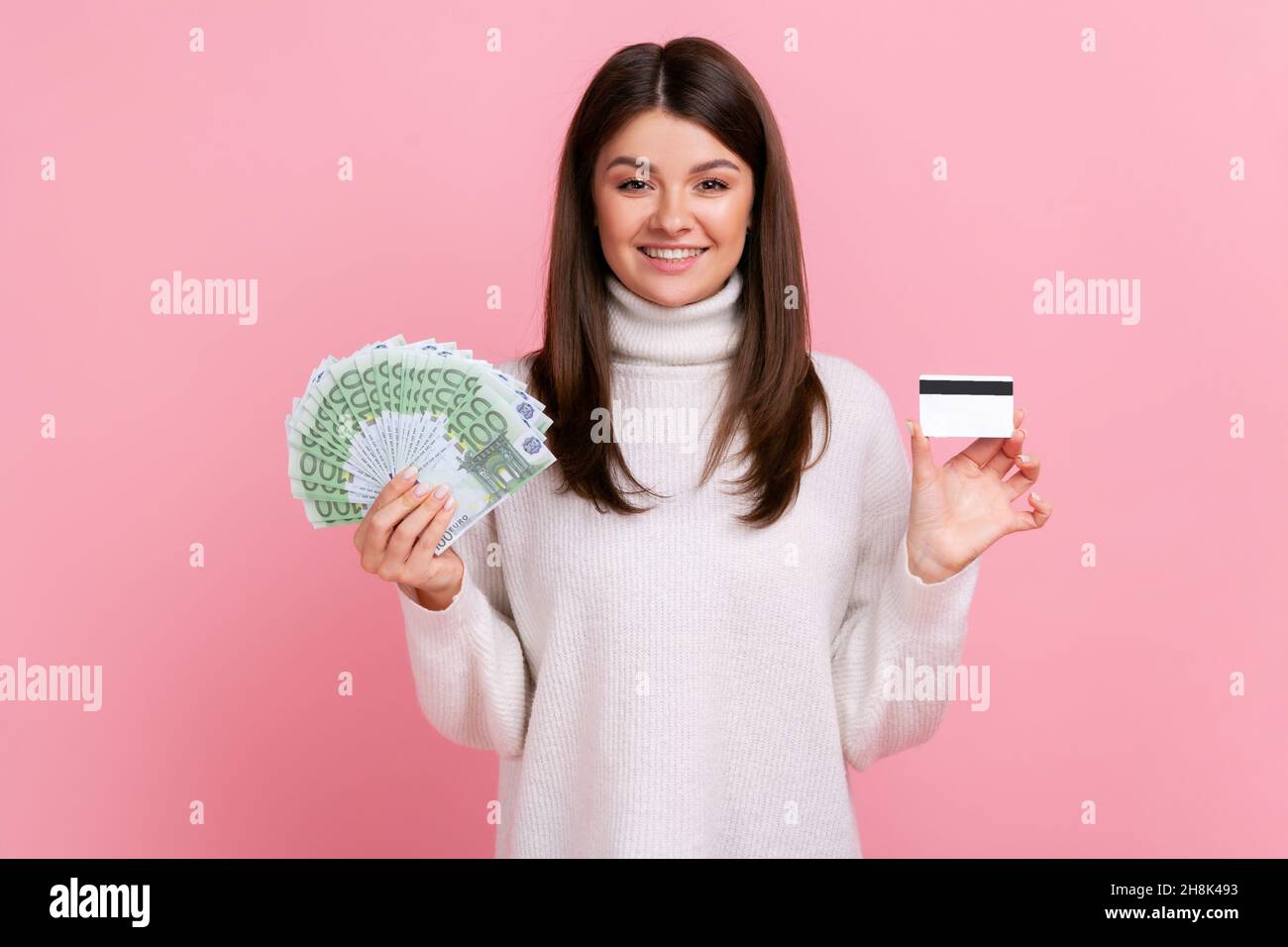 Femme souriante tenant une carte de crédit et grand fan de billets en euros, banque, gagnant une grosse somme d'argent, portant blanc pull de style décontracté.Studio d'intérieur isolé sur fond rose. Banque D'Images