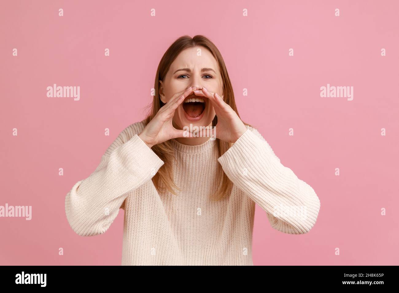 Portrait d'une jeune femme blonde adulte agressive debout, tenant les bras près de la large bouche ouverte et criant fort, portant un chandail blanc.Studio d'intérieur isolé sur fond rose. Banque D'Images