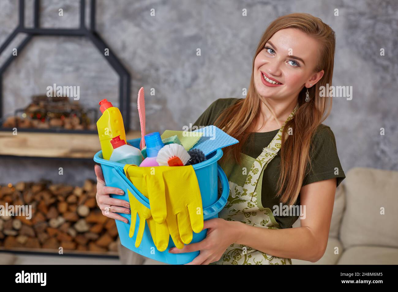 Une femme de ménage est debout dans la salle de séjour, tenant un seau bleu rempli de produits chimiques et d'installations pour ranger dans sa main Banque D'Images