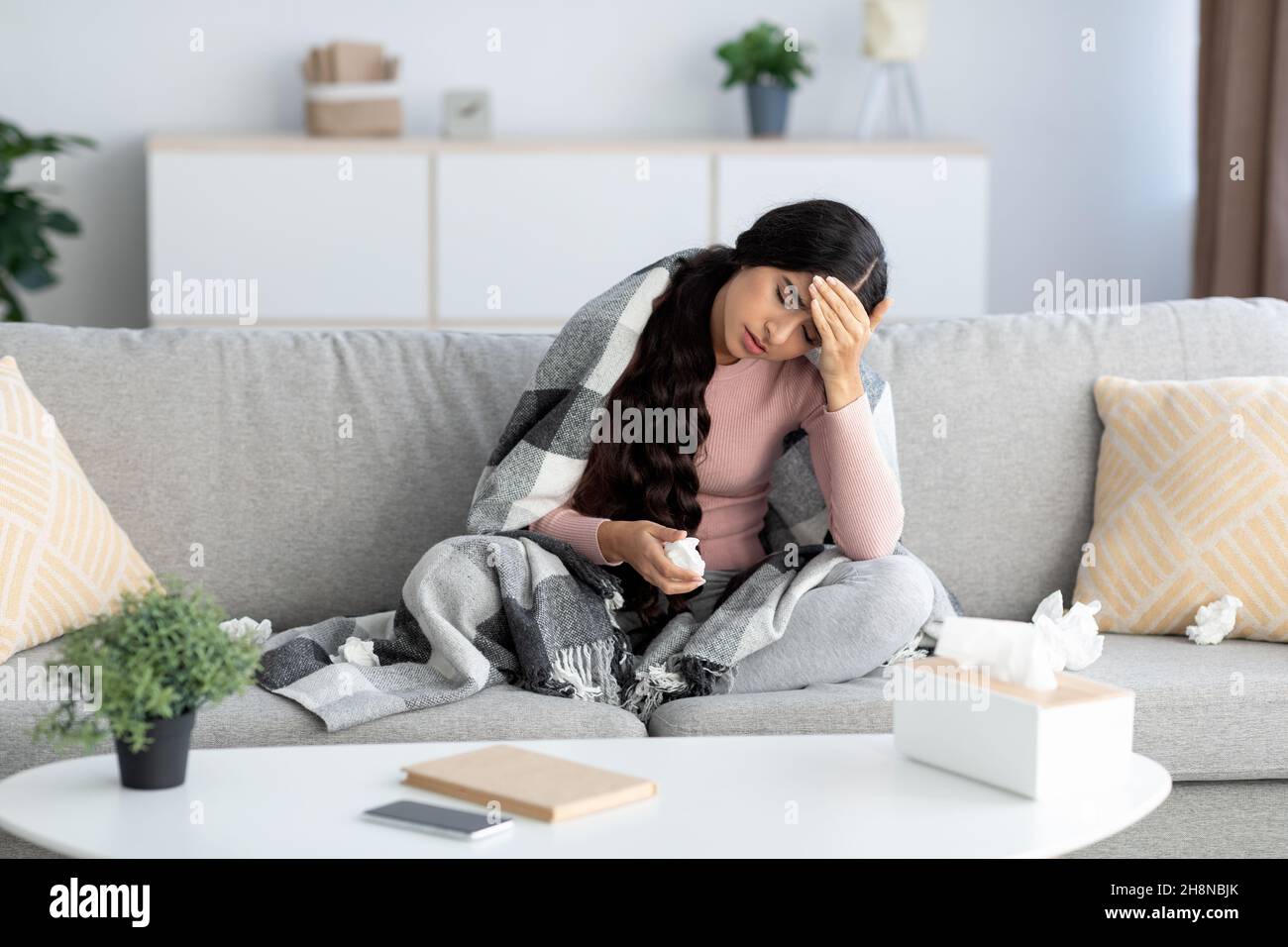 Jeune femme indienne en couverture, souffre de la grippe et des maux de tête, est assise sur un canapé à l'intérieur du salon Banque D'Images