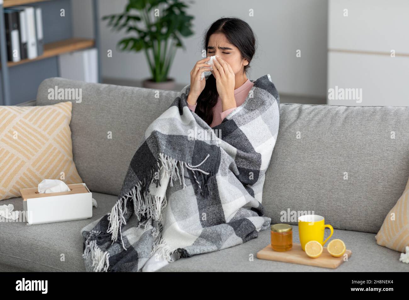 Une jeune femme indienne malheureuse éternue dans une serviette, souffle le nez, souffre de fièvre, de grippe sur le canapé dans la chambre avec du thé Banque D'Images