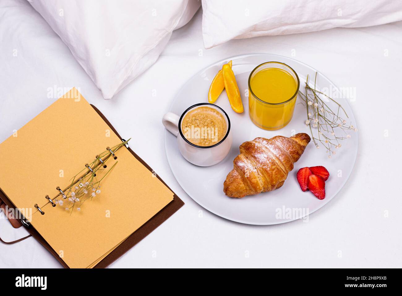 Carnet de croquis en papier, plaque en céramique avec croissant, café et jus d'orange sur le lit.Petit déjeuner romantique dans le lit. Banque D'Images