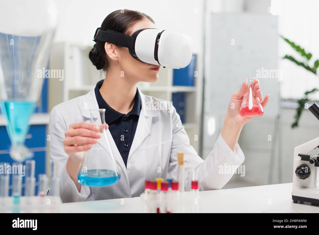 Un scientifique en réalité virtuelle tient des flacons près du microscope dans un laboratoire Banque D'Images