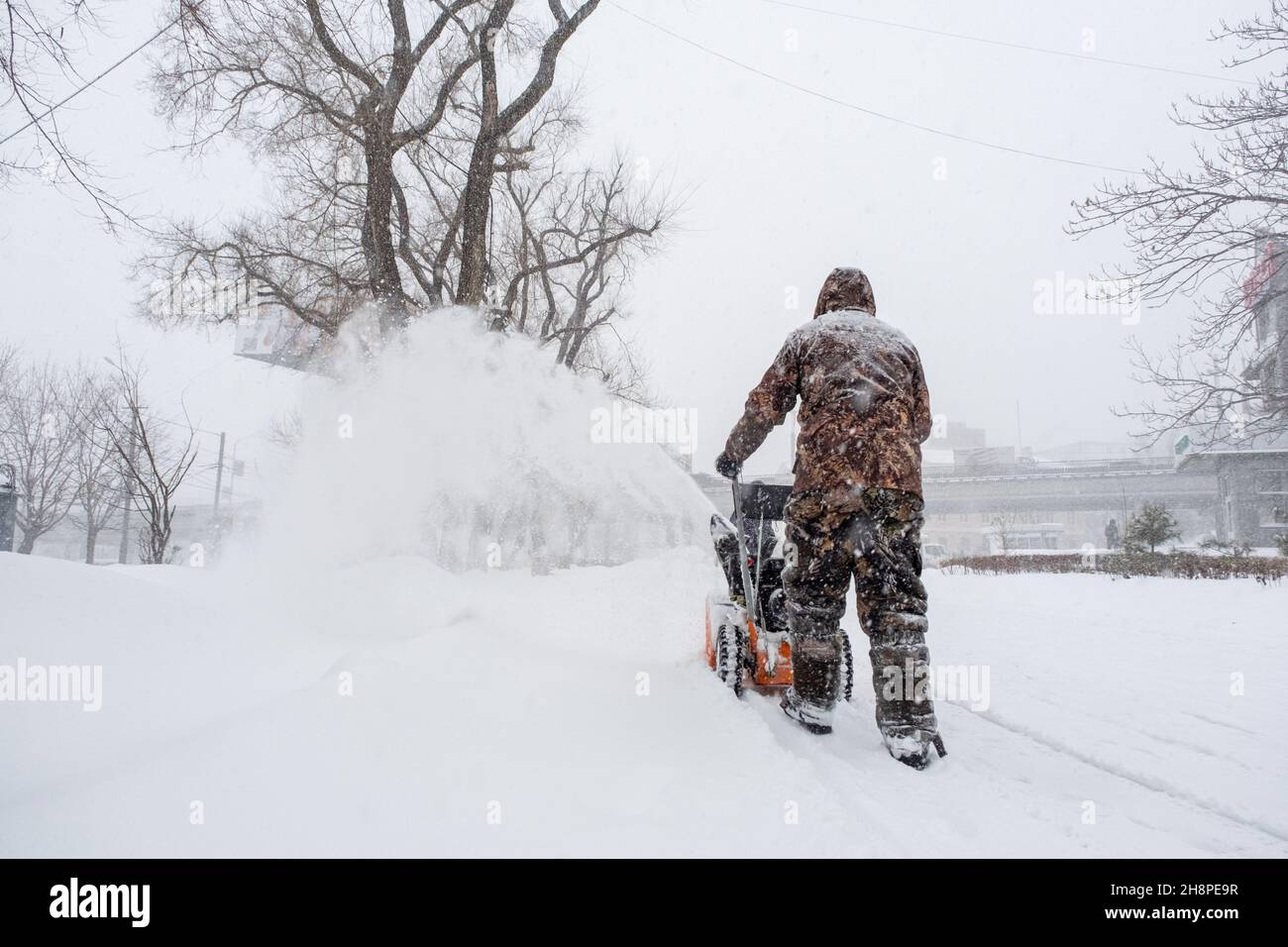 Un homme en camouflage marche dans la neige et déblayage la neige avec un chasse-neige à essence.Vue arrière. Banque D'Images