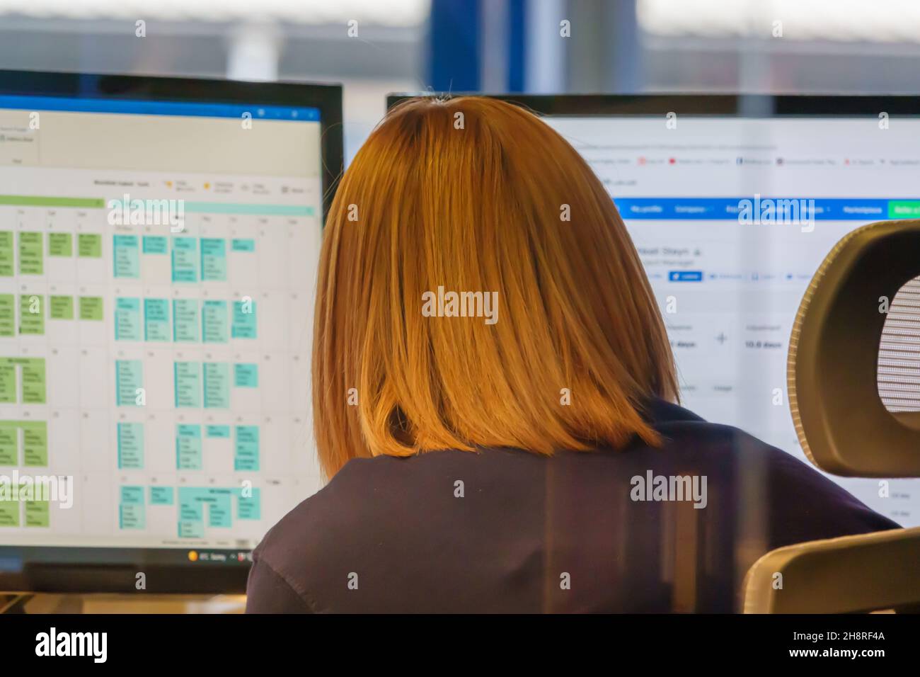 Une employée de bureau aux cheveux rouges devant les moniteurs de PC avec un calendrier de bureau flou à l'écran Banque D'Images