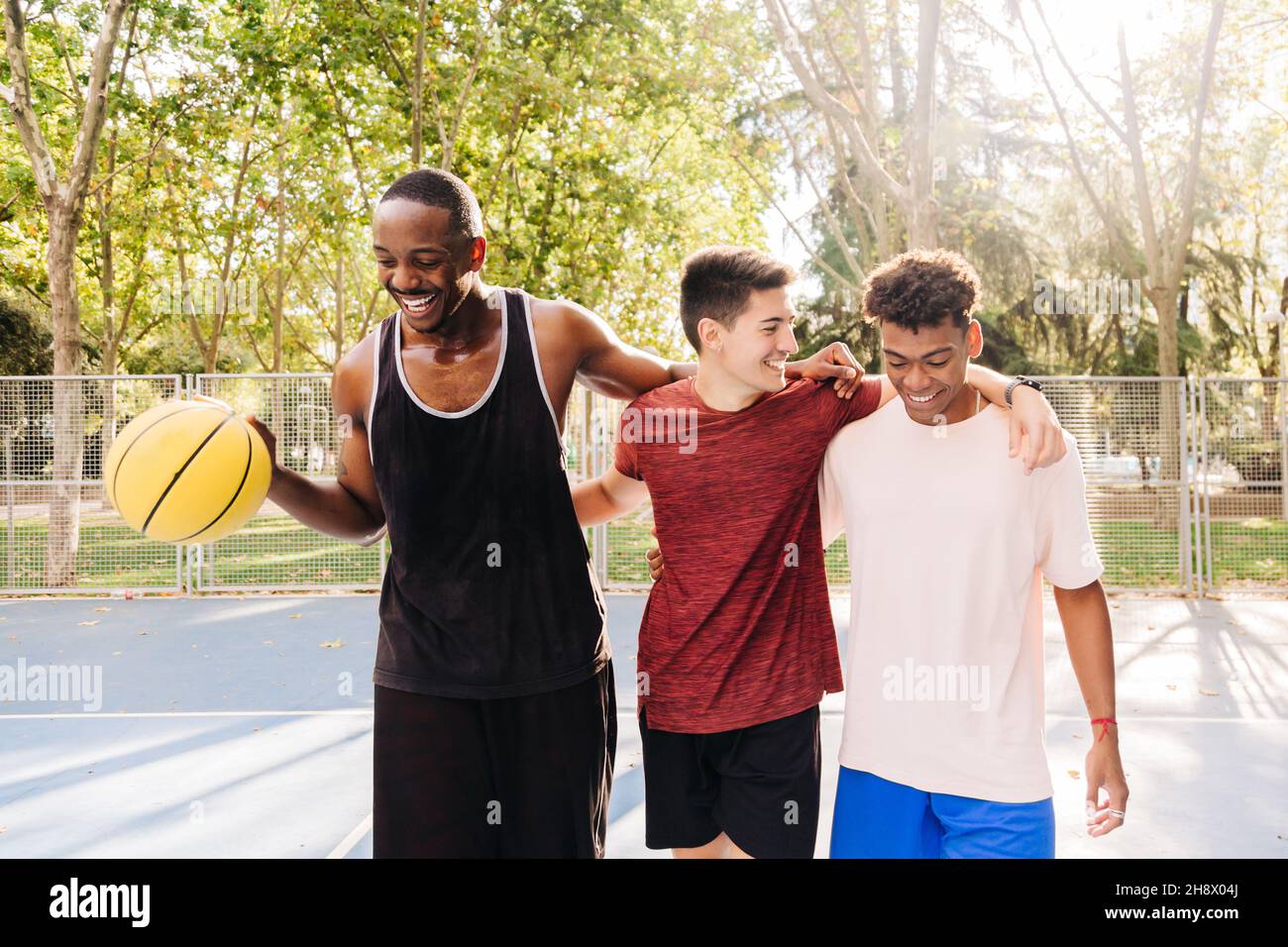 Des athlètes masculins gais et multiraciaux dans des vêtements de sport avec une balle de basket-ball jaune riant parler et embrassant tout en se tenant près de la clôture dans le stade Banque D'Images