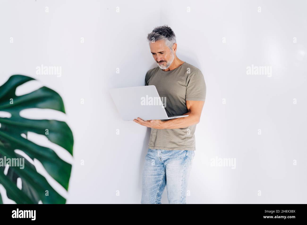 Homme âgé barbu regardant attentivement l'écran tout en se tenant contre un mur blanc et en parcourant un ordinateur portable à la main dans une pièce lumineuse Banque D'Images