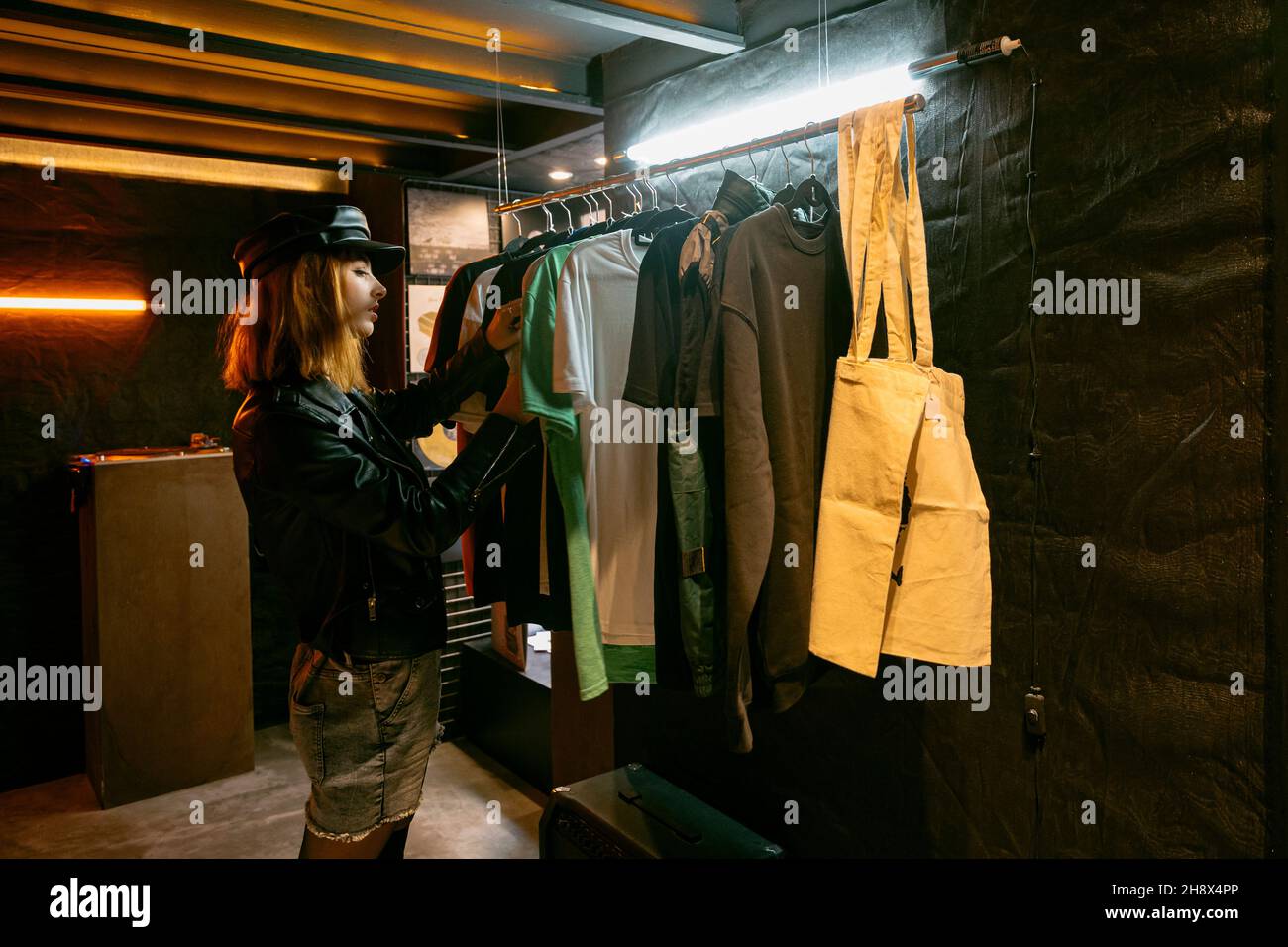 Vue latérale d'une femme sérieuse debout près d'un support en métal avec différents t-shirts tout en choisissant des vêtements en magasin avec une faible lumière Banque D'Images Vue latérale d'une femme sérieuse debout près d'un support en métal avec différents t-shirts tout en choisissant des vêtements en magasin avec une faible lumière Banque D'Images