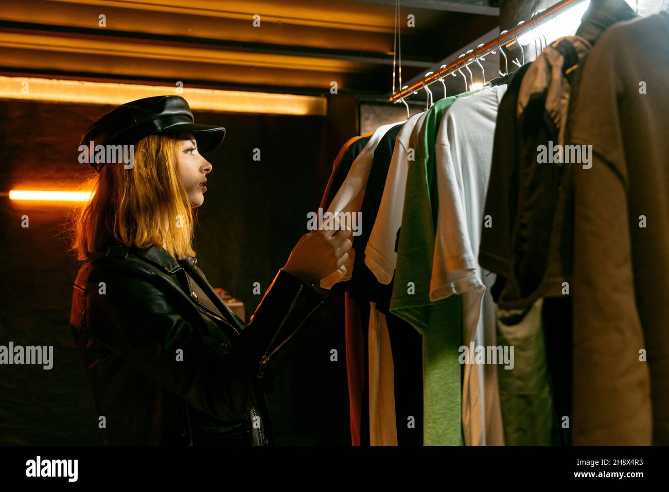 Vue latérale d'une femme sérieuse debout près d'un support en métal avec différents t-shirts tout en choisissant des vêtements en magasin avec une faible lumière Banque D'Images Vue latérale d'une femme sérieuse debout près d'un support en métal avec différents t-shirts tout en choisissant des vêtements en magasin avec une faible lumière Banque D'Images