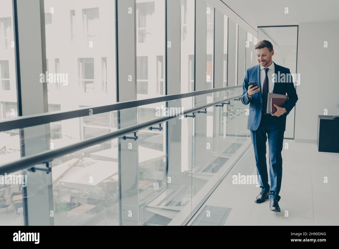 Souriant redhead homme employé de bureau en costume utilisant le téléphone portable tout en se tenant dans le bureau moderne Banque D'Images