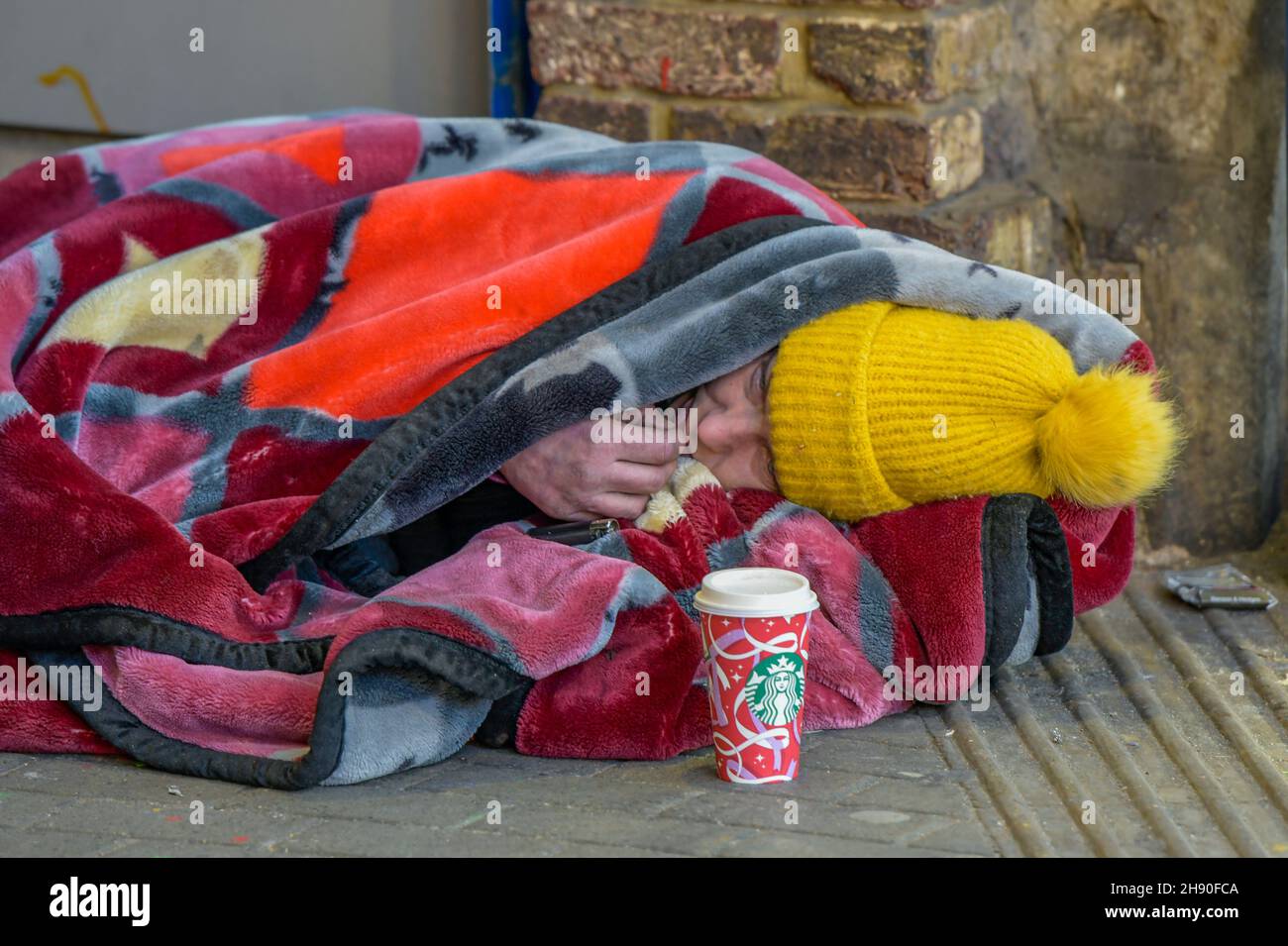 personne sans-abri enveloppée dans des couvertures portant un chapeau laineux avec une tasse de café starbucks dans une tasse de noël dormant difficilement sous un métro ou une arche. Banque D'Images