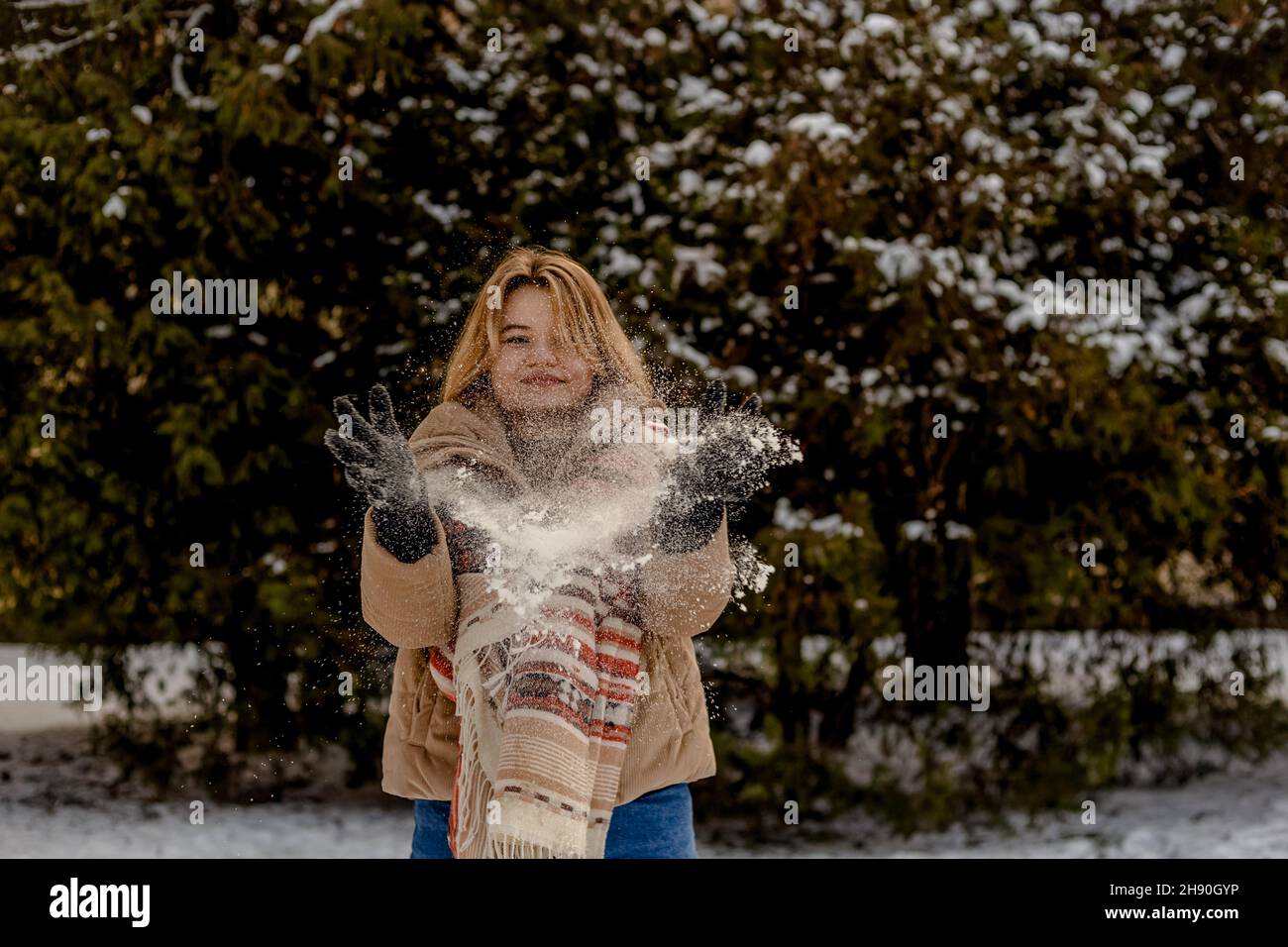 La jeune femme blonde en blouson et foulard beige jette de la neige avec ses mains.Arrière-plan des arbres de noël dans la neige.Saison d'hiver. Banque D'Images