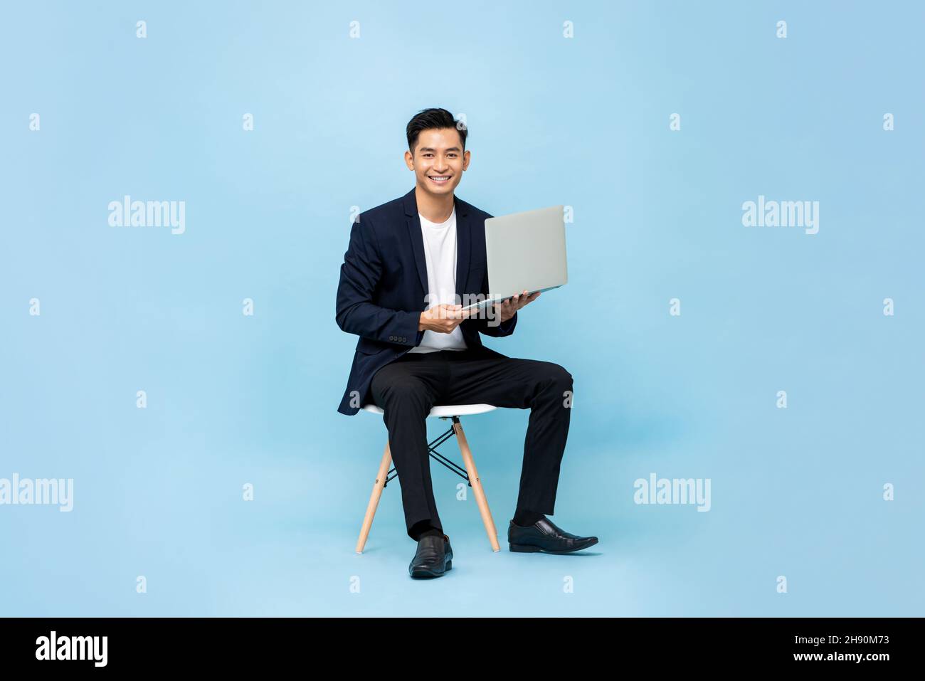 Portrait d'un jeune homme d'affaires souriant et charmant d'Asie du Sud-est avec ordinateur portable assis sur une chaise sur fond de studio bleu clair Banque D'Images