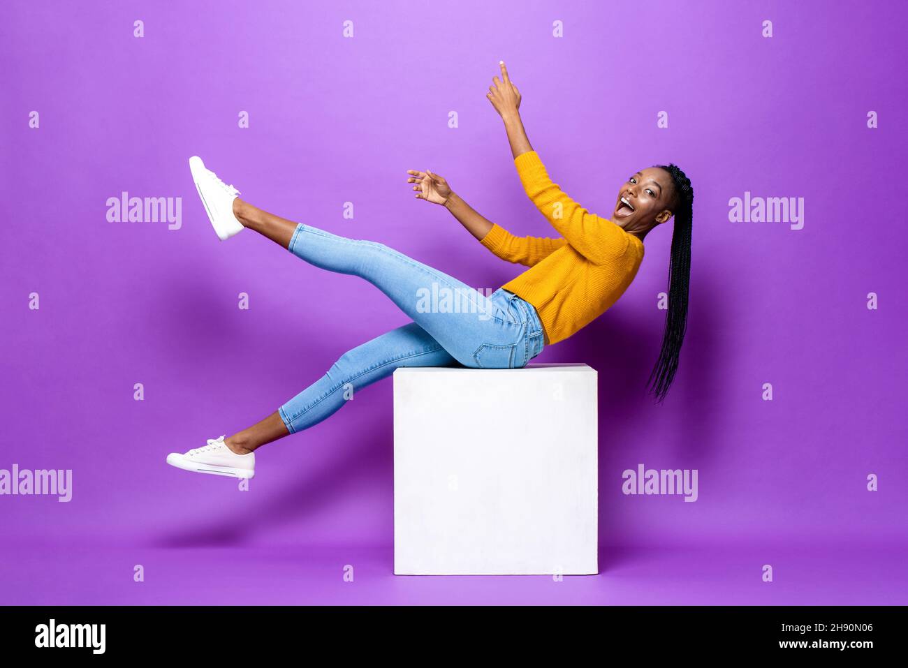 Jeune femme afro-américaine souriante, couchée sur un tabouret levant les mains et les jambes dans un arrière-plan isolé de couleur violet studio Banque D'Images