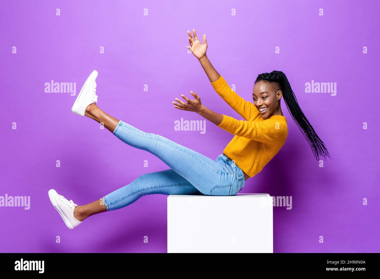 Jeune femme afro-américaine souriante, couchée sur un tabouret levant les mains et les jambes dans un arrière-plan isolé de couleur violet studio Banque D'Images
