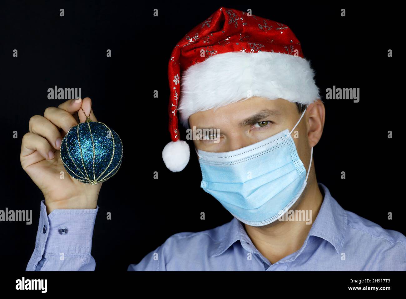 Fête de Noël, portrait d'homme en masque et chapeau de père Noël avec une balle de jouet bleue à la main.Concept de décoration de maison ou de bureau pour la nouvelle année Banque D'Images