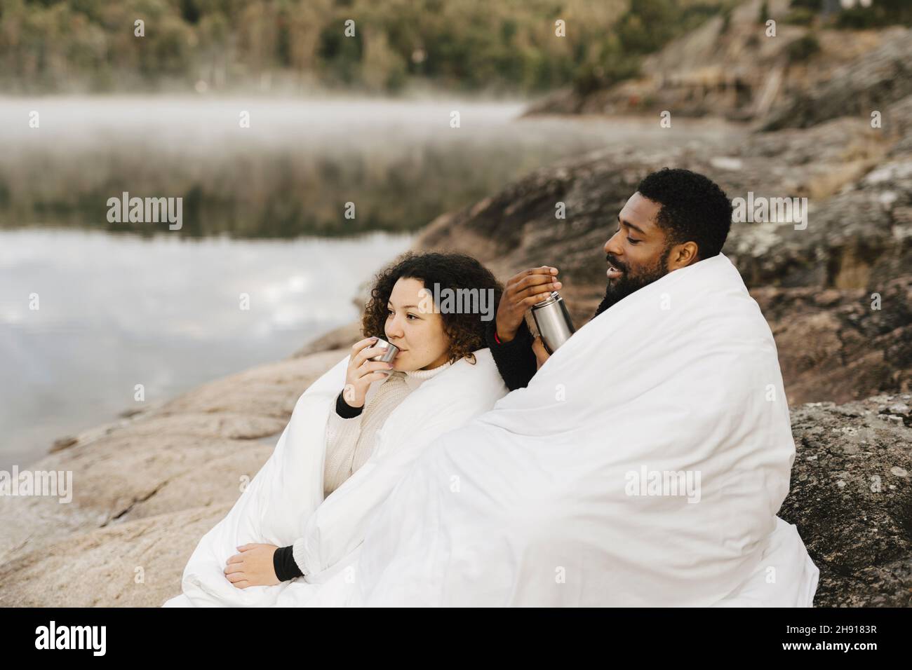 Femme enveloppée dans une couverture avec un petit ami tout en buvant au bord du lac Banque D'Images