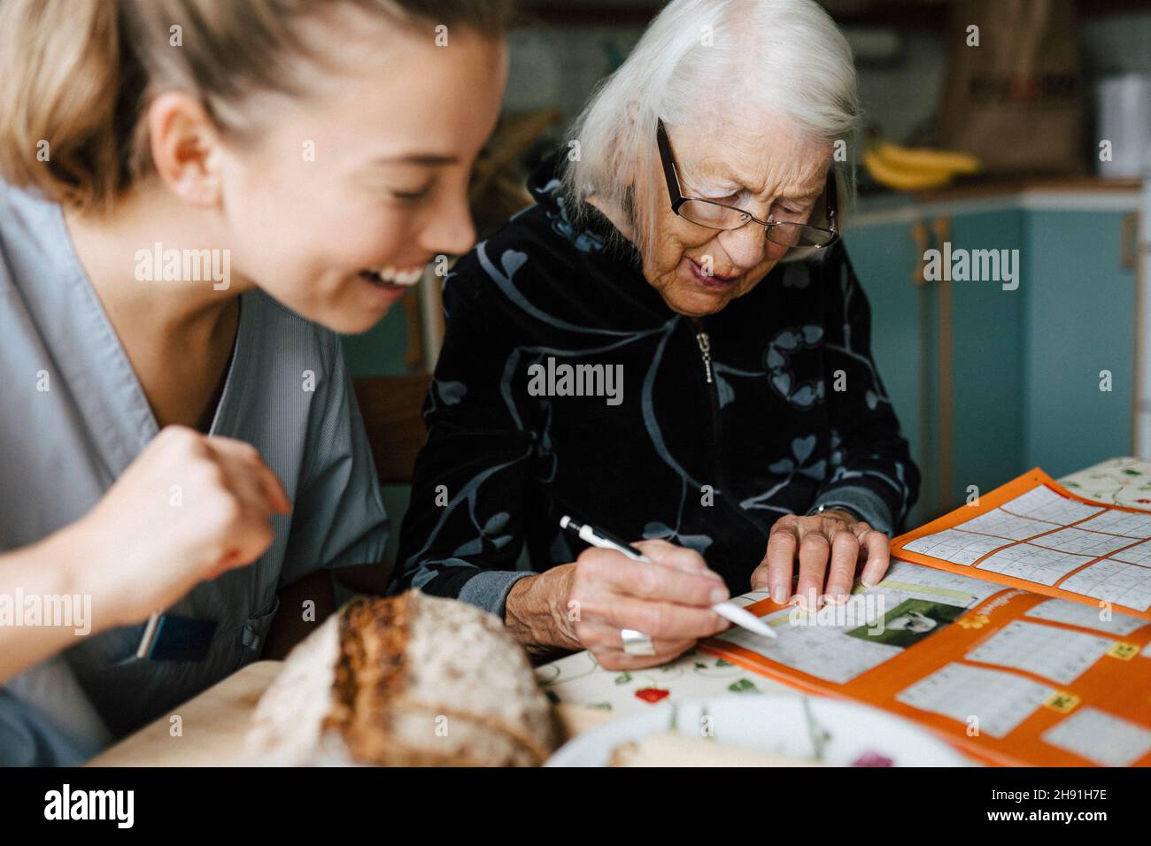Femme souriante qui s'occupe de la santé pendant que la femme âgée résolvant le casse-tête des mots croisés dans la cuisine Banque D'Images