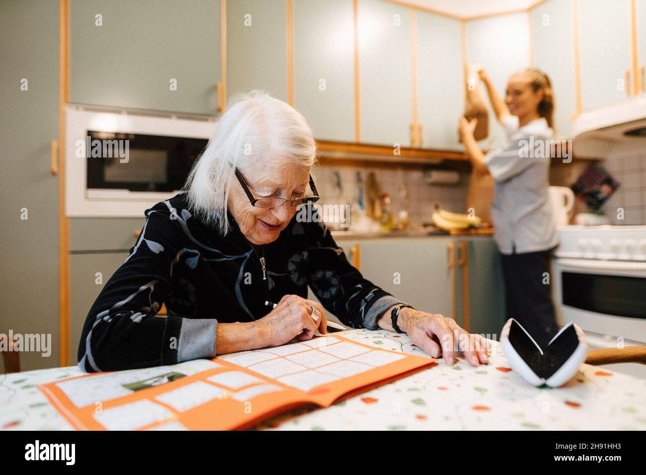 Femme senior résolvant le sudoku tandis que l'infirmière féminine fait des corvées dans la cuisine Banque D'Images