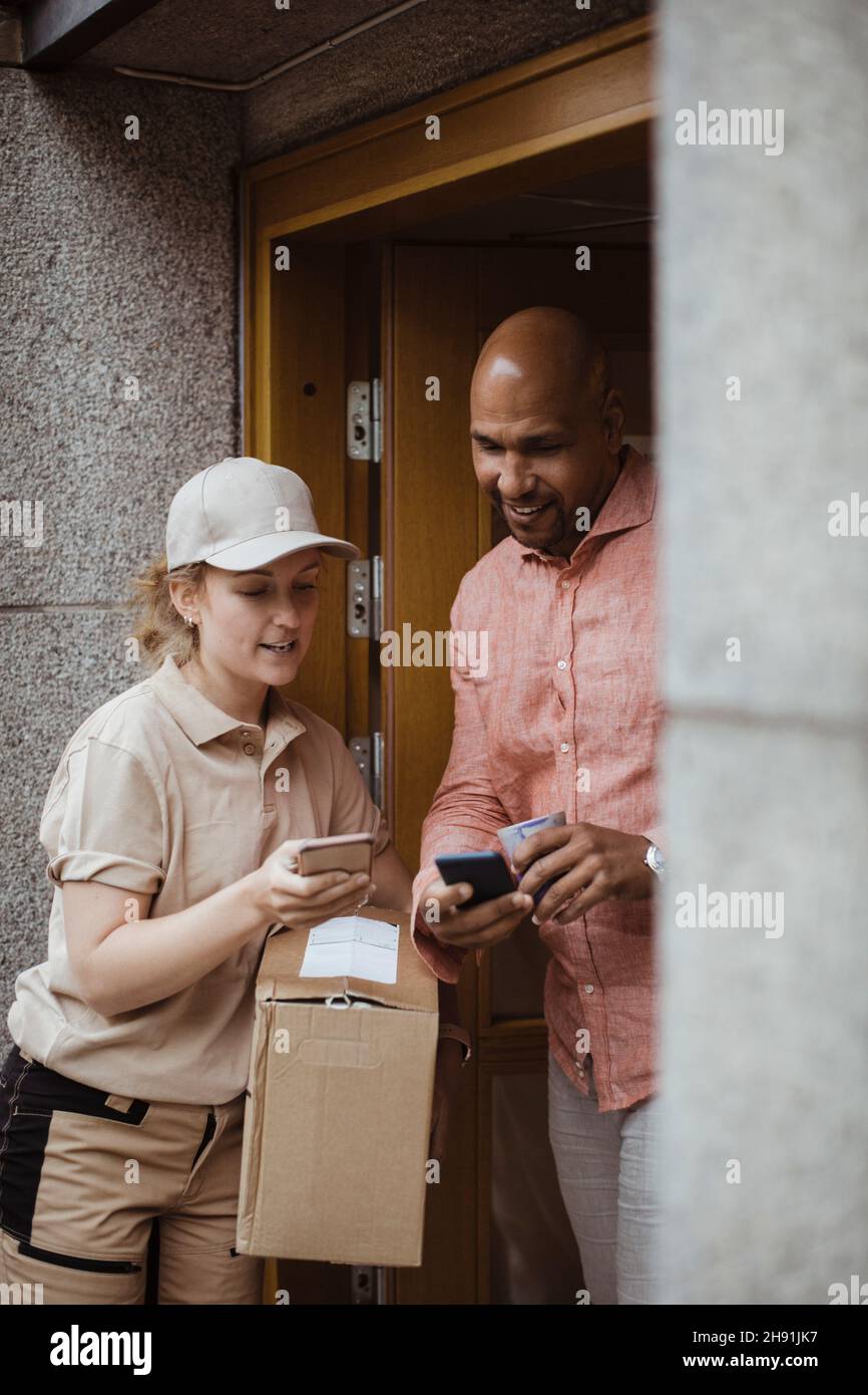 Jeune femme de livraison et client masculin utilisant des smartphones à la porte Banque D'Images