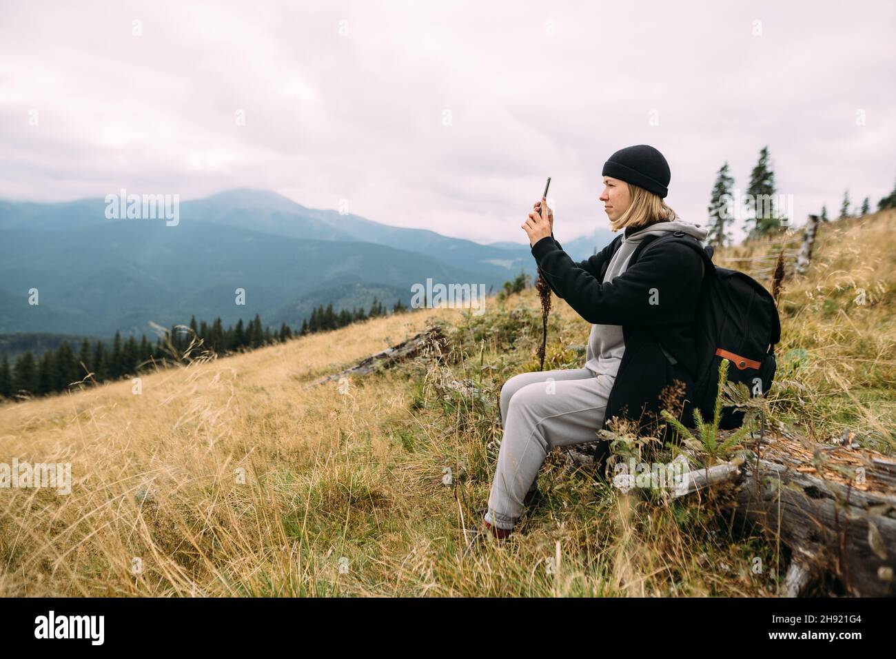 Une heureuse touriste s'assoit au sommet d'une montagne pendant un arrêt, prenant une photo sur un smartphone mobile, Blogger, en regardant les montagnes. Banque D'Images