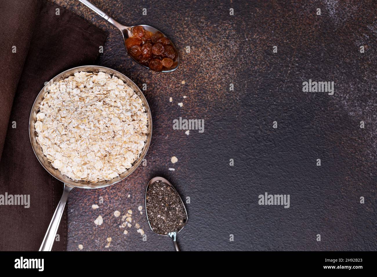 Ingrédients pour faire du porridge de flocons d'avoine.Composition alimentaire avec flocons d'avoine dans une casserole en métal, chia et raisins secs dans des cuillères sur un dos texturé brun foncé Banque D'Images