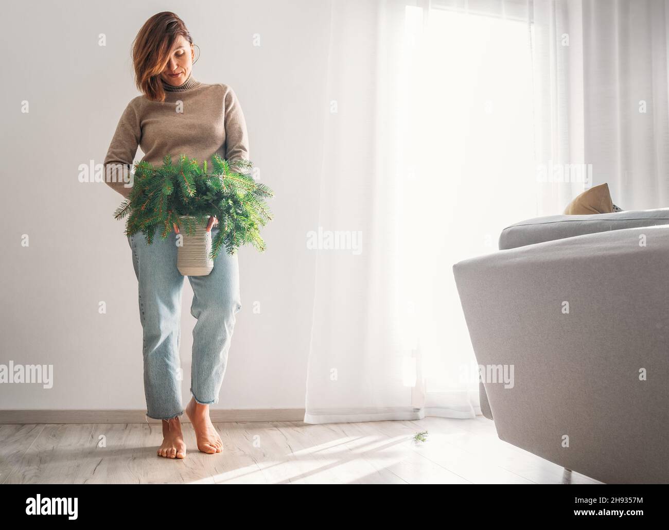 Une jeune femme vêtue chaud confortable chandail tenant le vase en argile rempli de branches vertes de sapin fraîchement coupé conifères près d'un mur blanc dans la maison liv Banque D'Images