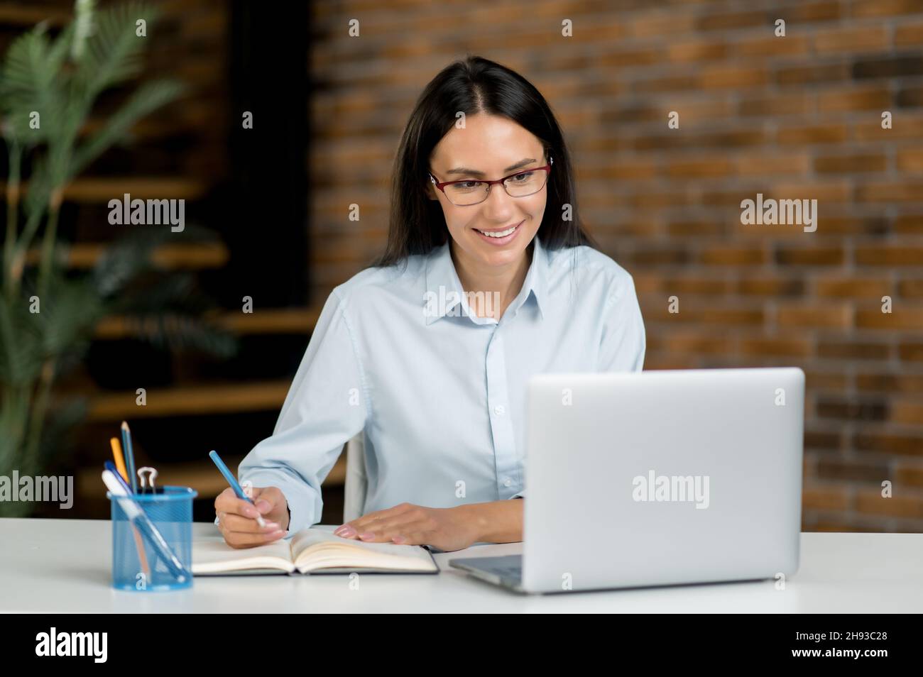 Jolie femme brune, élégante et réussie, de race blanche, s'assoit au bureau, étudie ou travaille sur un ordinateur portable, écoute une conférence en ligne, un séminaire en ligne, note des informations, communique par appel vidéo Banque D'Images