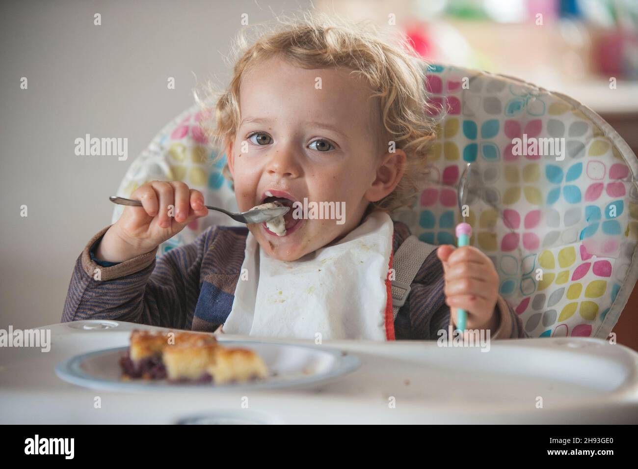 Une petite fille de 2 ans mange un peu de gâteau et de crème glacée comme collation de l'après-midi sur une chaise haute. Banque D'Images