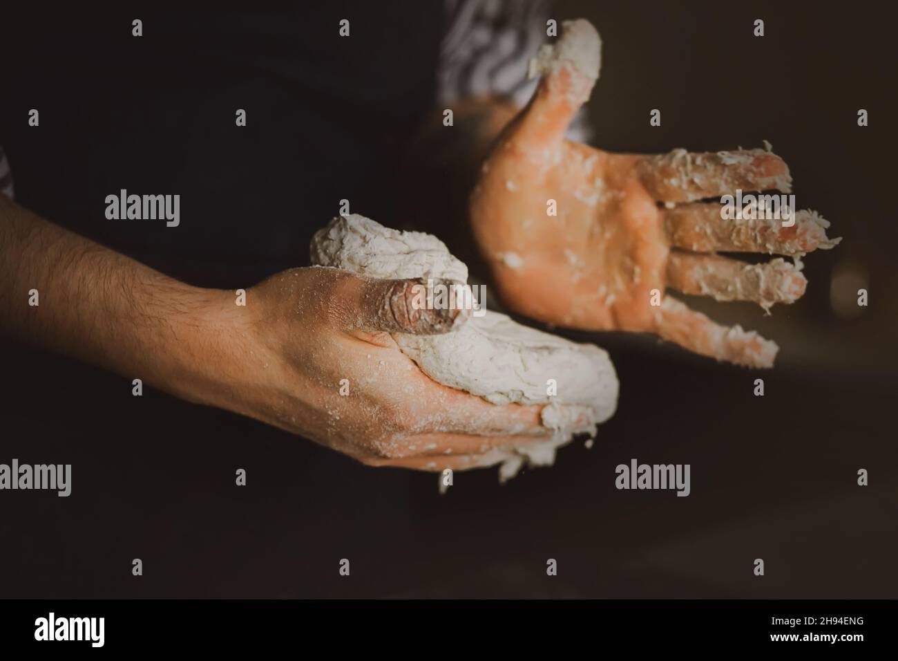 Un homme dans un tablier foncé pétrir de la pâte de blé fraîche avec ses mains dans la cuisine.Le processus de fabrication du pain et de la cuisson.Cuisine maison. Banque D'Images