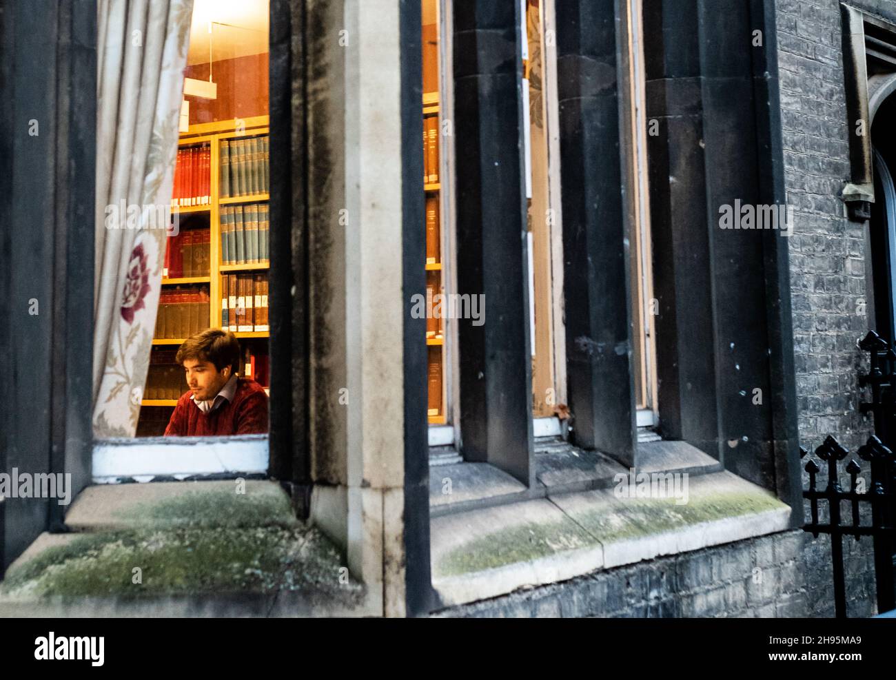 Un étudiant de premier cycle à l'Université de Cambridge étudie seul dans une chambre dans un ancien bâtiment avec des murs tapissés de livres dans une soirée froide de décembre.Après Banque D'Images