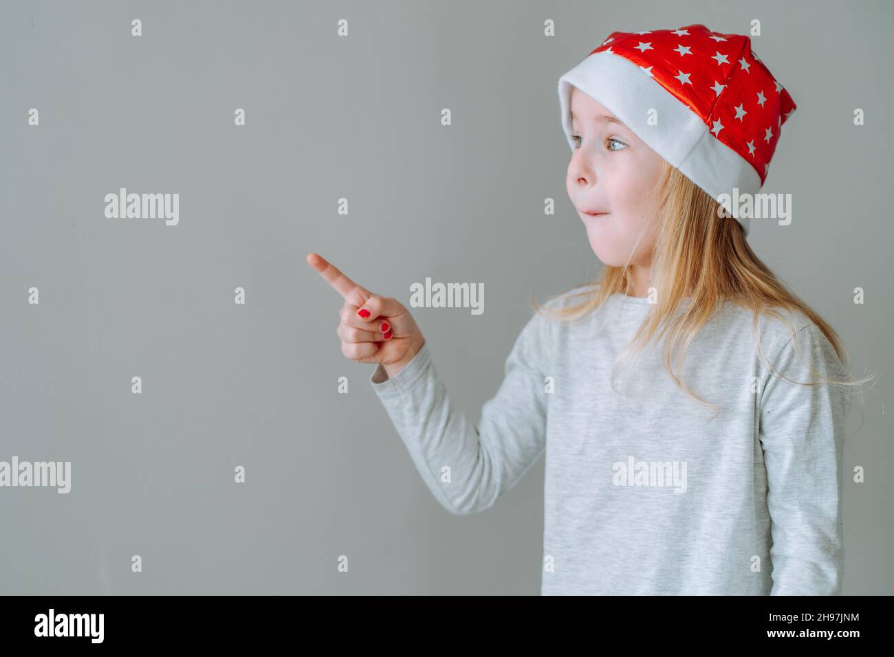 Une jeune fille portant un chapeau de père noël rouge pointe son doigt vers un espace vide à côté d'elle contre le mur gris neutre.CopySpace. Banque D'Images
