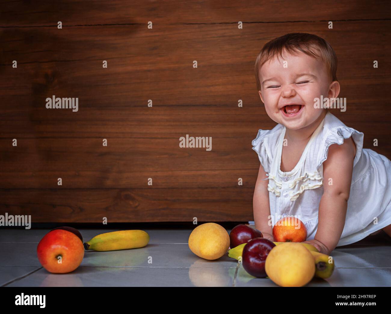 bonne petite fille mignonne rampant sur le sol avec des pommes de fruits, prunes, bananes, mangue sur un fond de bois Banque D'Images