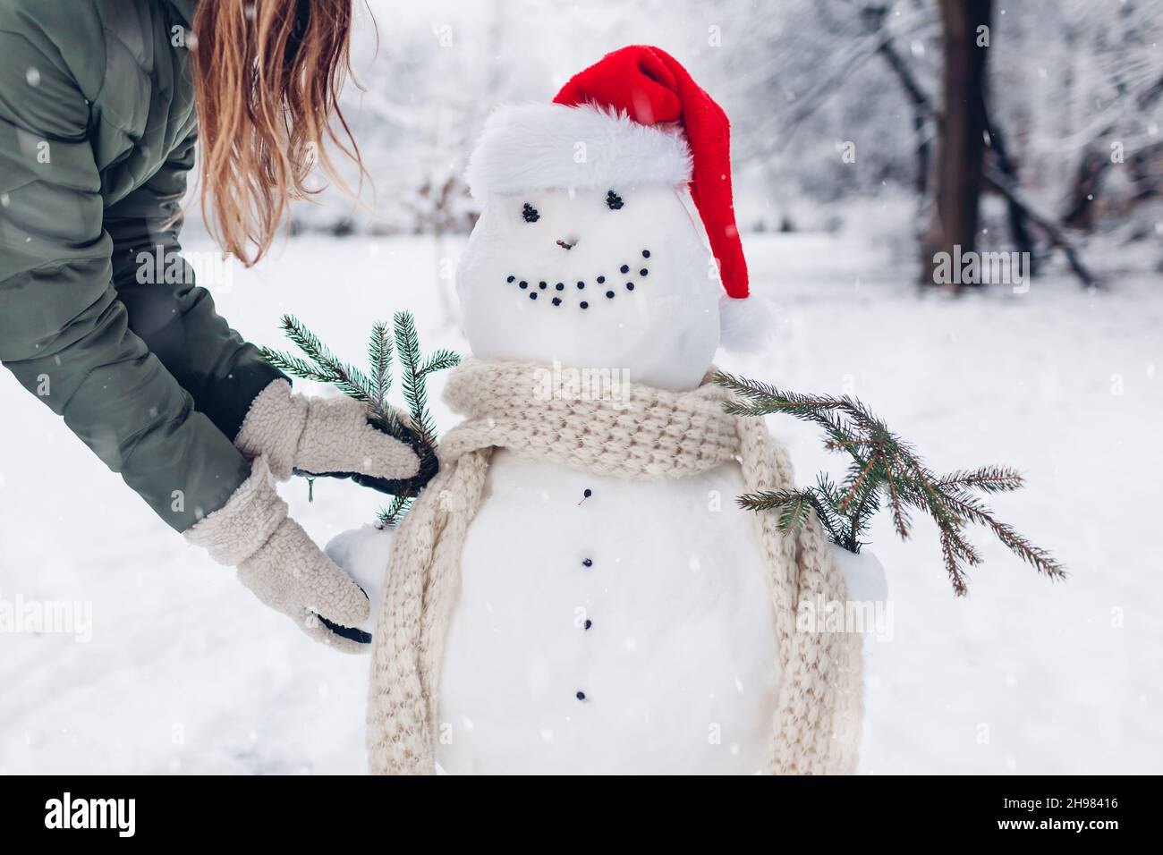 Fille fait bonhomme de neige dehors dans le parc d'hiver enneigé en le portant dans le chapeau de Santa, le foulard et la fixation des branches de sapin dans les mains.Activités de Noël Banque D'Images