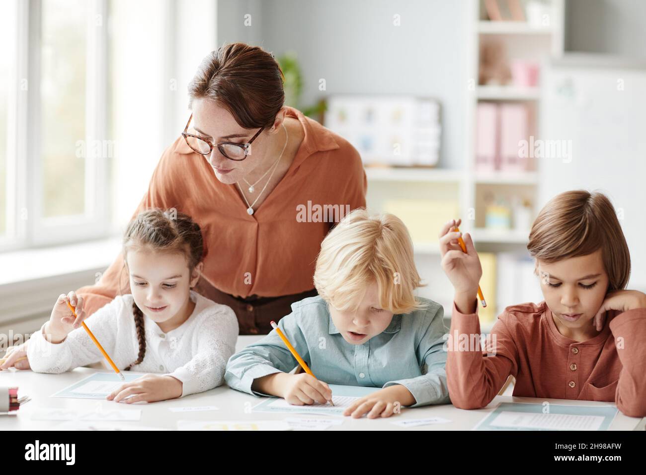 Jeune professeur de lunettes aidant les enfants à faire leurs devoirs à la table dans la salle de classe Banque D'Images