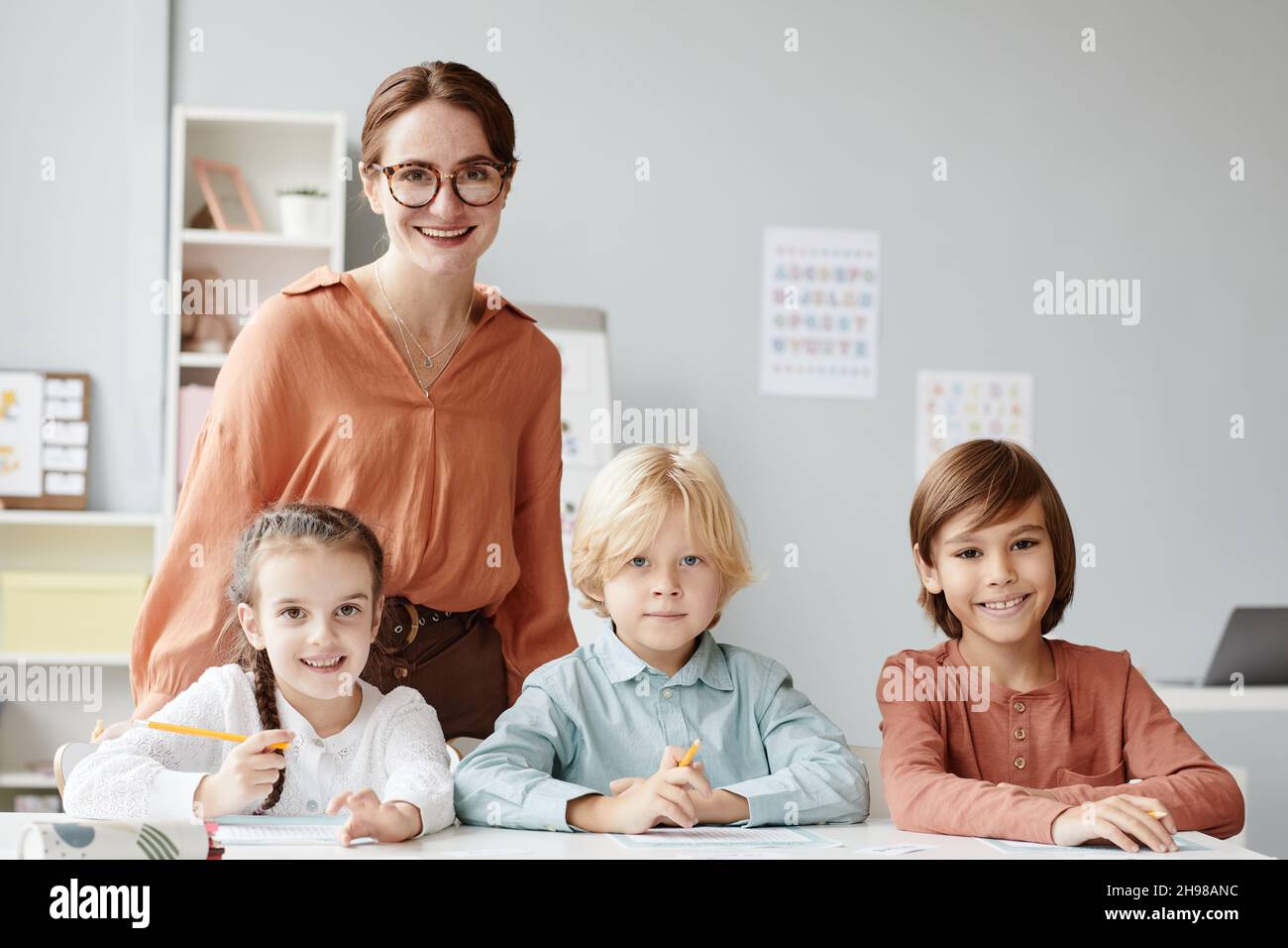 Portrait d'un jeune enseignant en lunettes souriant à la caméra avec un groupe d'enfants assis à la table de la classe Banque D'Images