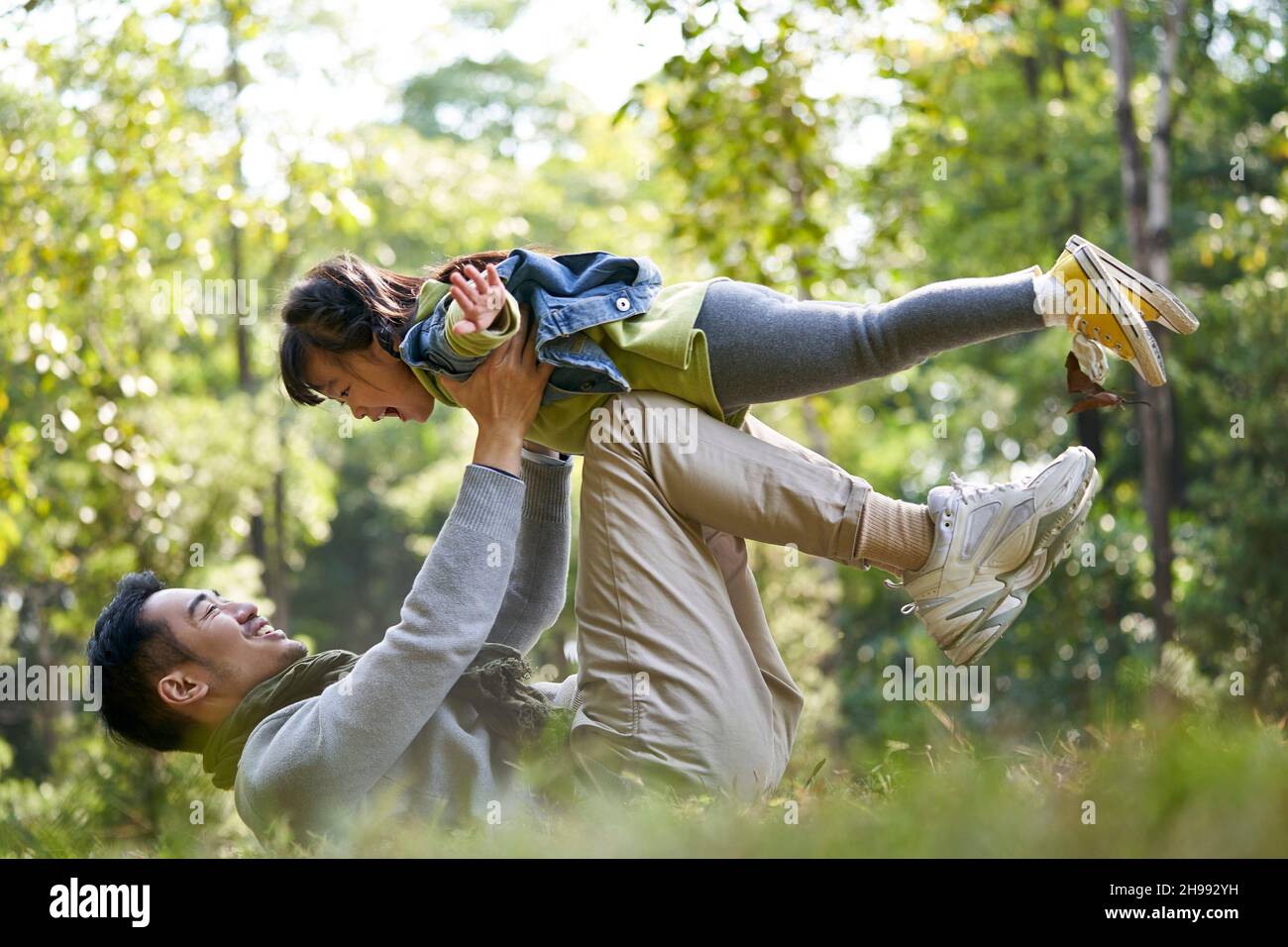 père asiatique allongé sur le dos sur l'herbe ayant un bon moment de soulever la fille dans les airs à l'extérieur dans le parc de la ville Banque D'Images