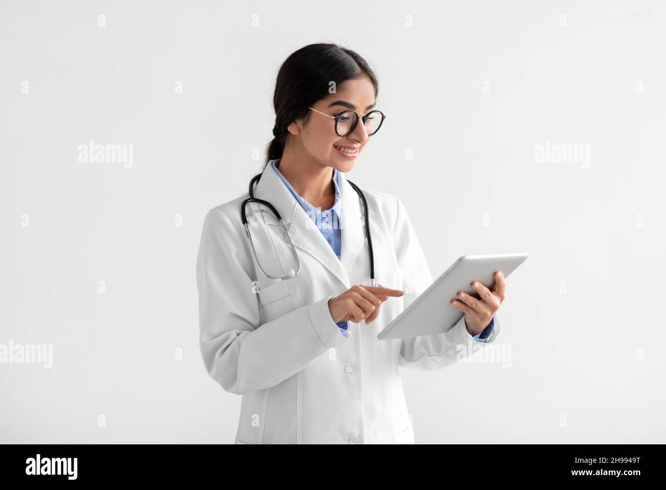 Bonne jeune femme indienne médecin en manteau blanc et lunettes travaillant sur tablette Banque D'Images