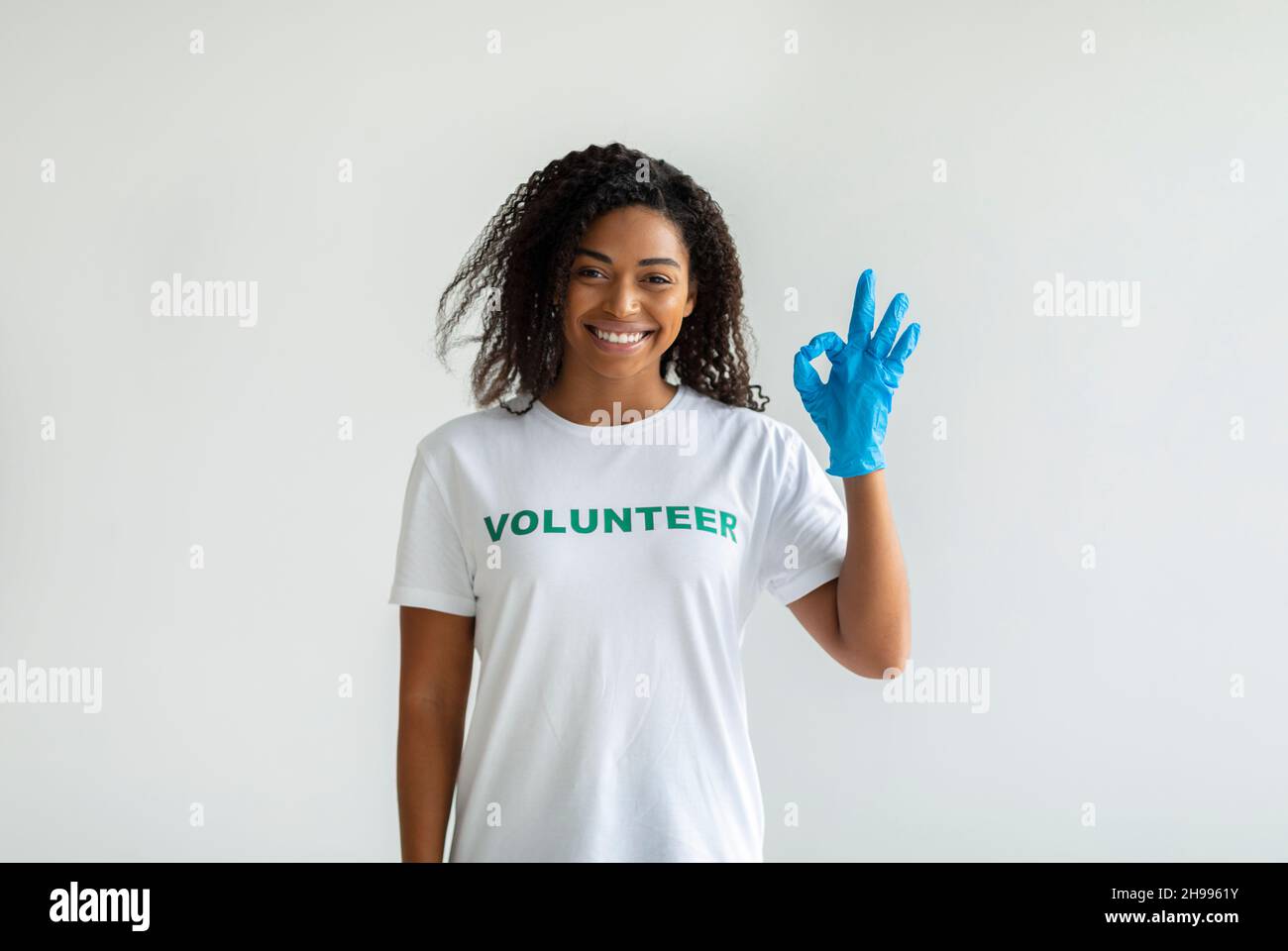 Concept de grâce de charité.Bonne femme volontaire dans des gants montrant le geste OK et souriant à l'appareil photo, mur léger Banque D'Images