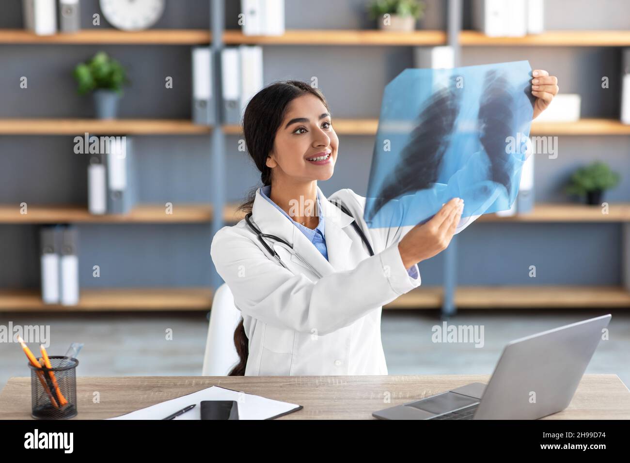 Bonne jeune femme indienne médecin en manteau blanc regarder la radiographie des poumons image dans l'intérieur de bureau de clinique Banque D'Images