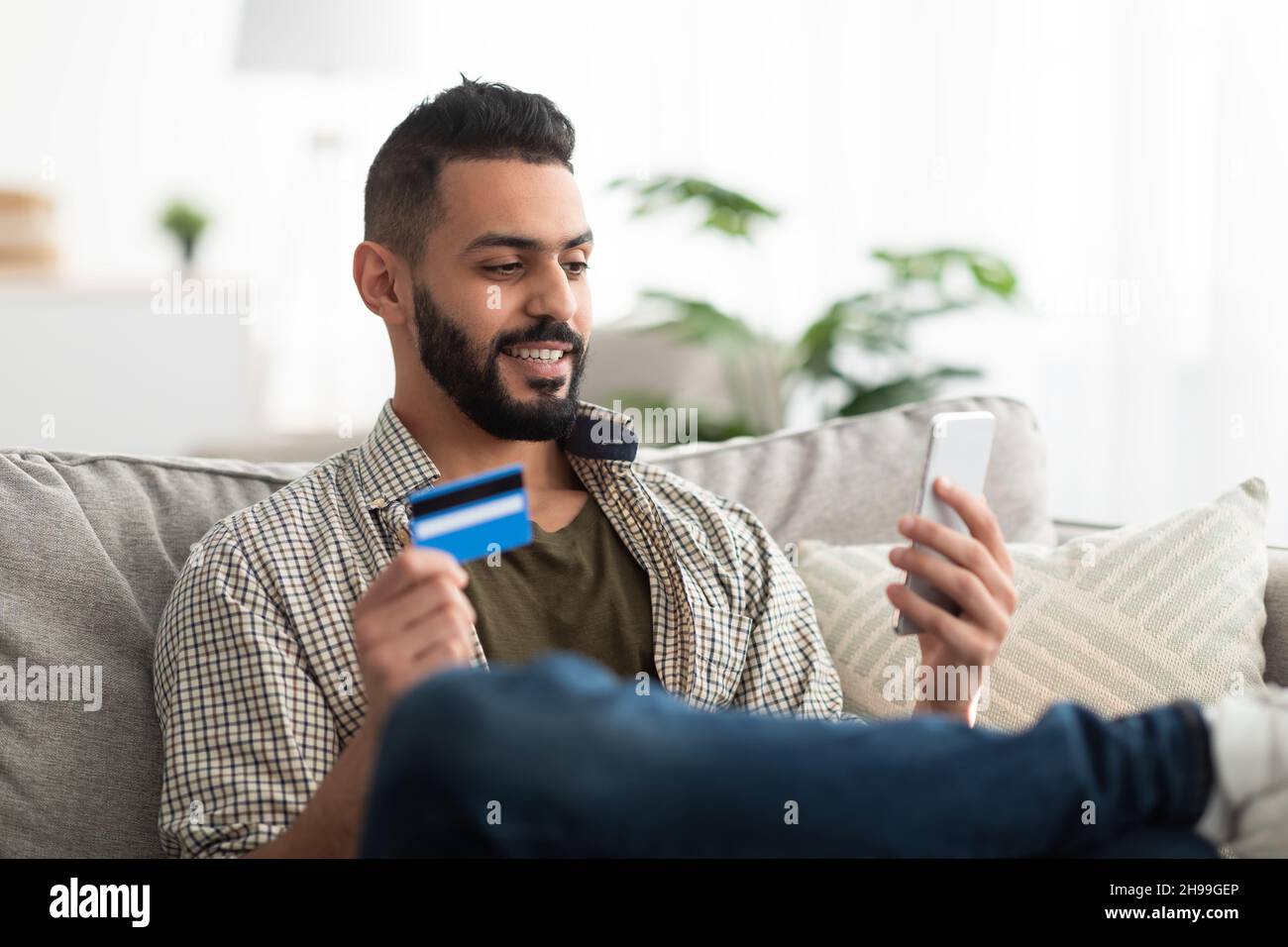 Jeune homme arabe attrayant utilisant un téléphone mobile et une carte de crédit pour faire des achats en ligne ou des opérations bancaires à distance à la maison Banque D'Images
