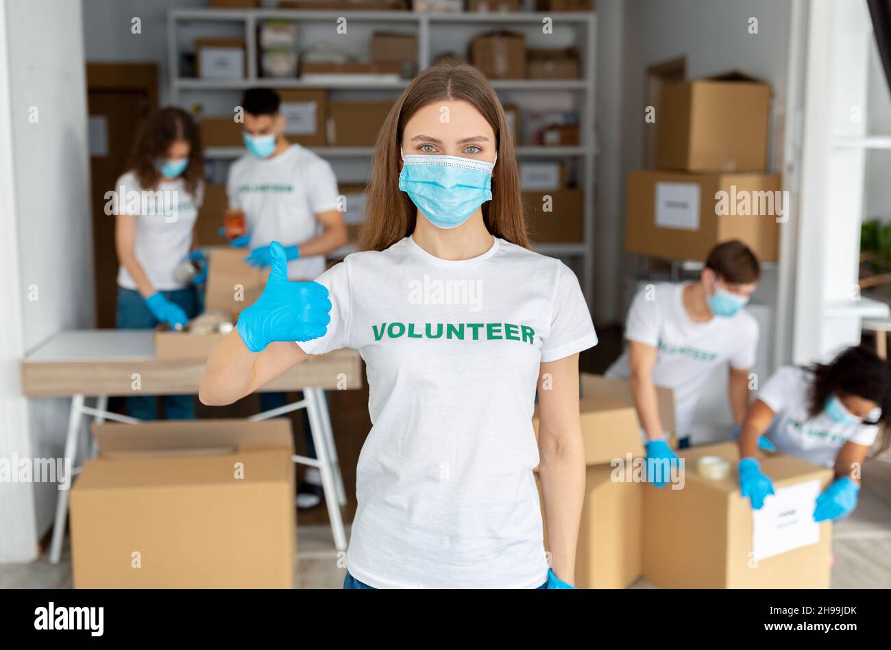 Militante féminine portant un masque médical, travaillant au centre de bénévoles, montrant le pouce vers le haut à la caméra Banque D'Images